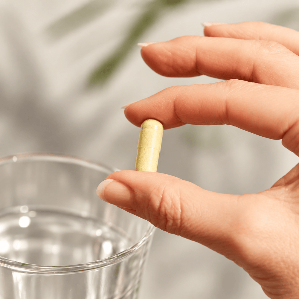 A close-up of a hand holding a single berberine capsule pill above a clear glass of water, with a softly blurred background.