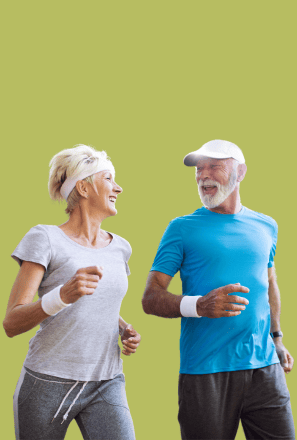 Older man and woman jogging together, smiling and looking at each other, wearing athletic clothing against a plain background.