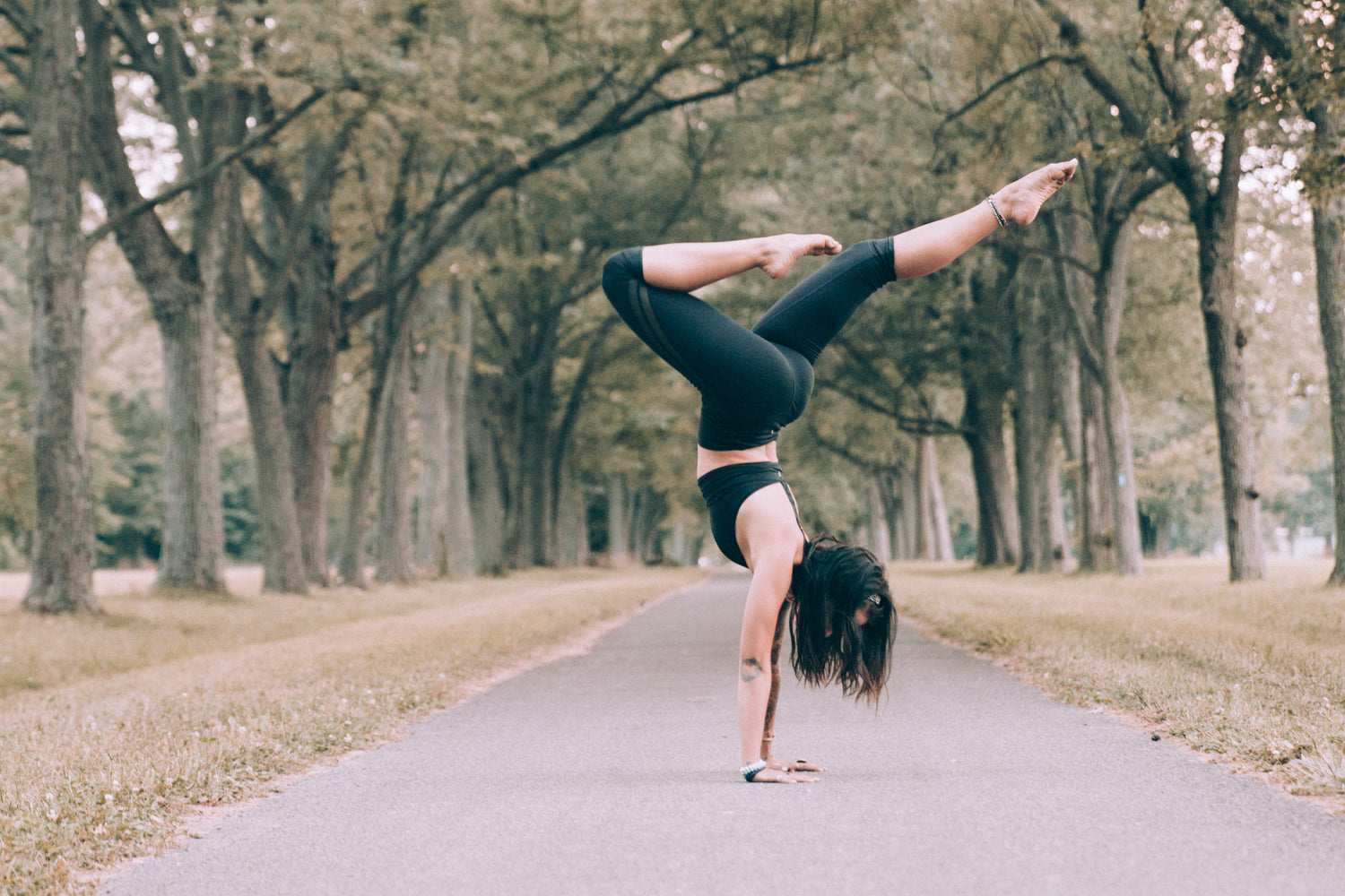 Person performing a one-handed handstand yoga pose on a paved path lined with tall trees, wearing black athletic clothing, with legs extended in a split position and hair hanging downward.